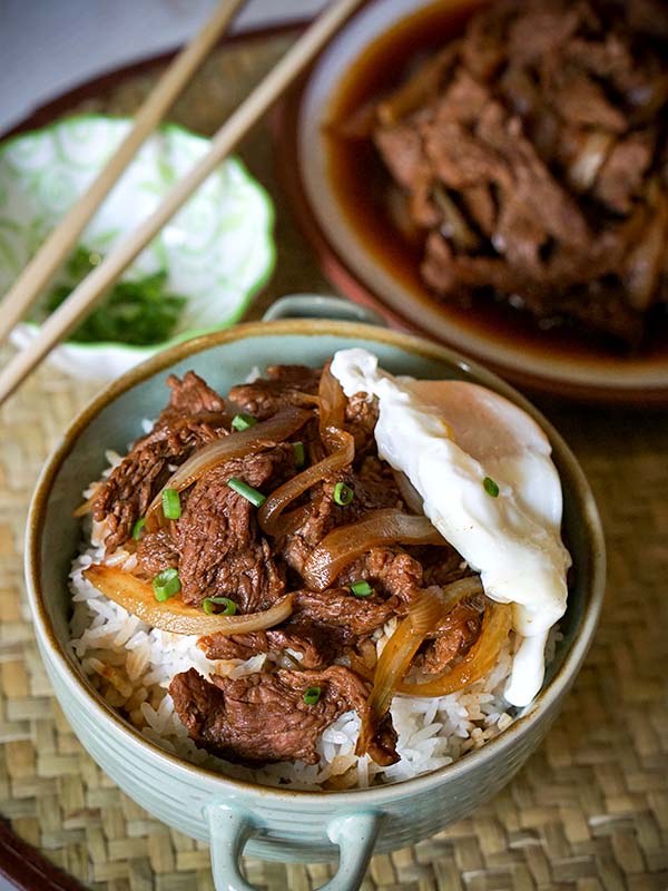 beef with onions on top of rice with egg in a bowl on top of table